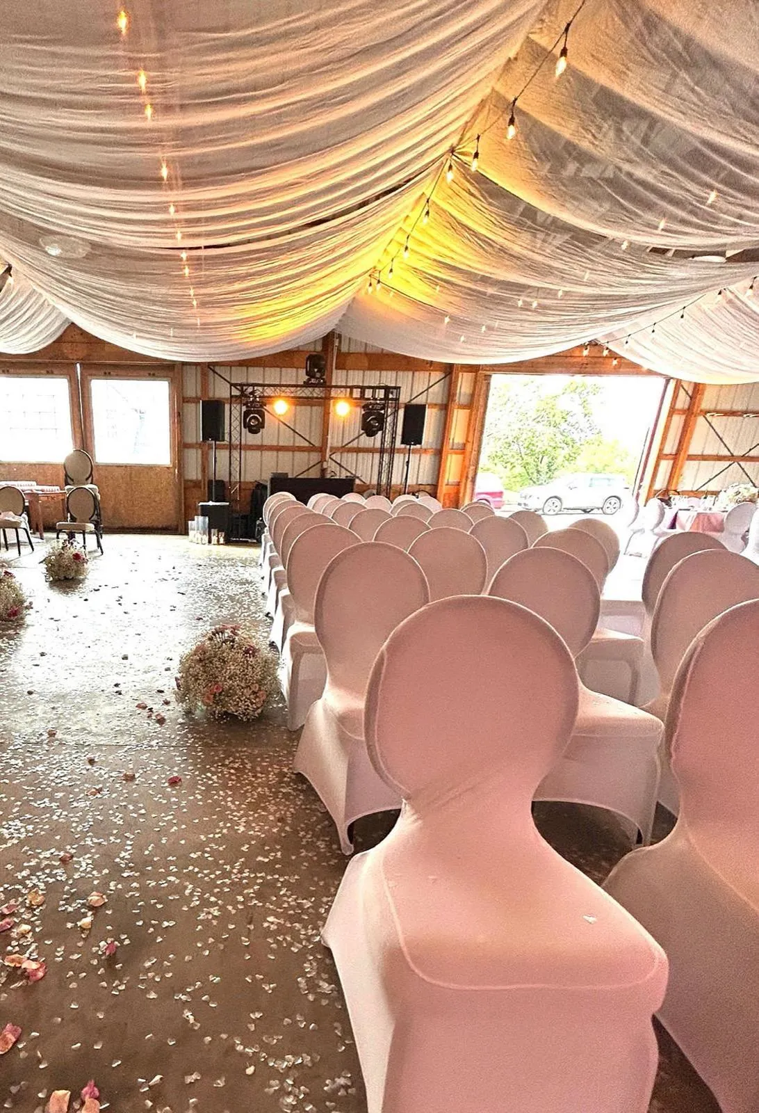 Inside the barn: a draped fabric ceiling, warm string lights, blush chair covers and petals scattered along the aisle.