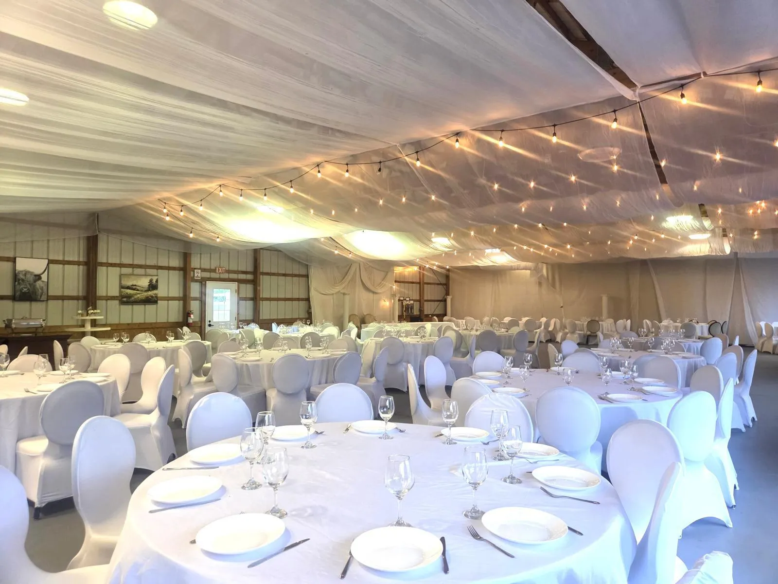 An all-white reception: round tables set with glassware beneath a draped ceiling crossed with strings of warm lights.