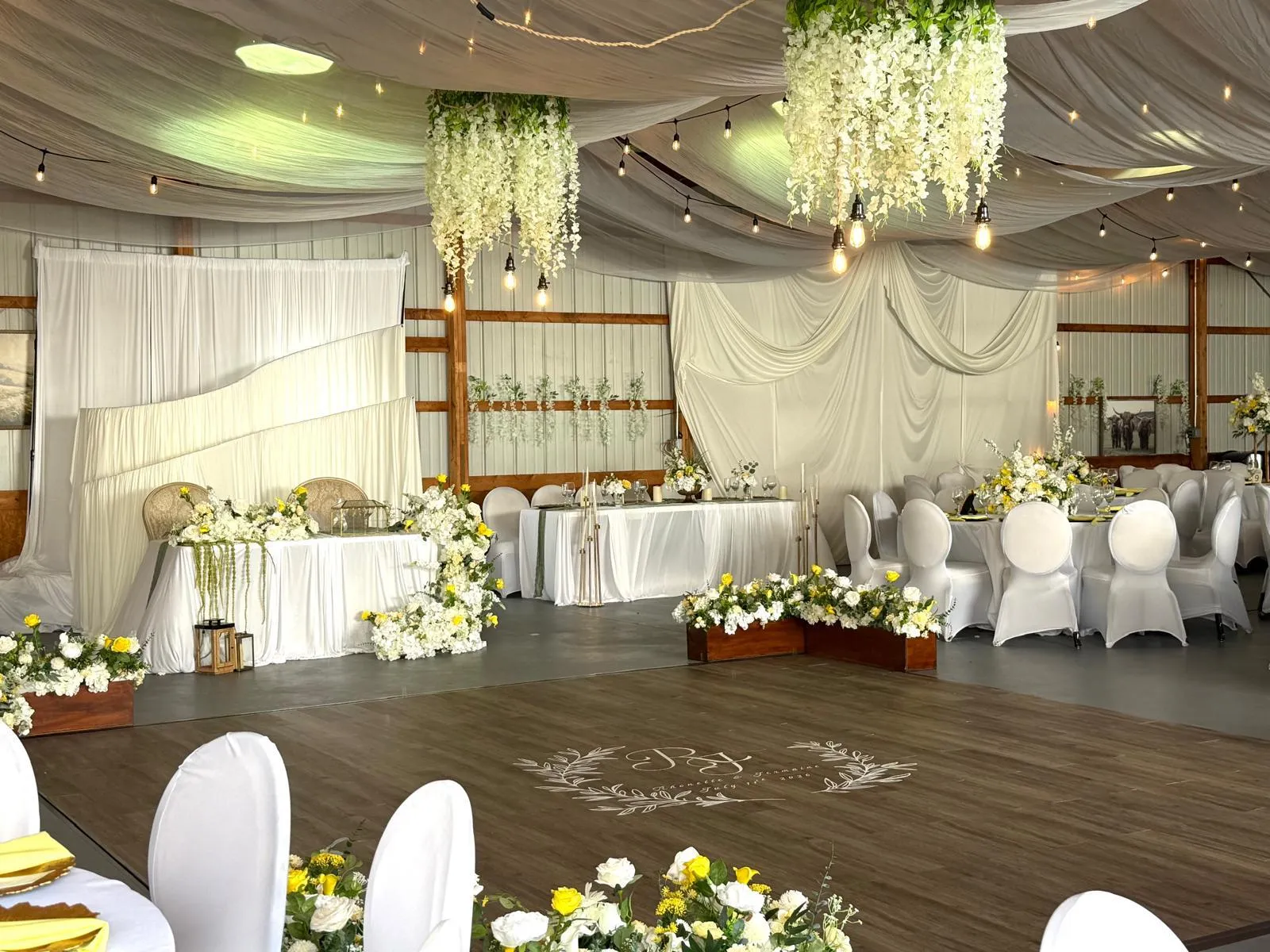 A reception room with a wooden dance floor at its center, hanging white wisteria, draped ceiling and a sweetheart table banked with yellow and white flowers.