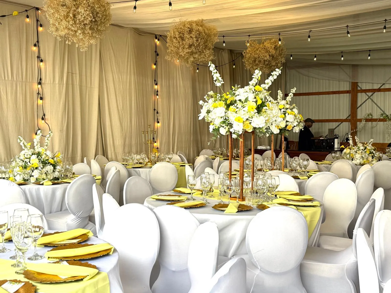 Round tables in yellow and white with gold chargers, tall floral centerpieces on brass stands and dried baby's breath spheres overhead.