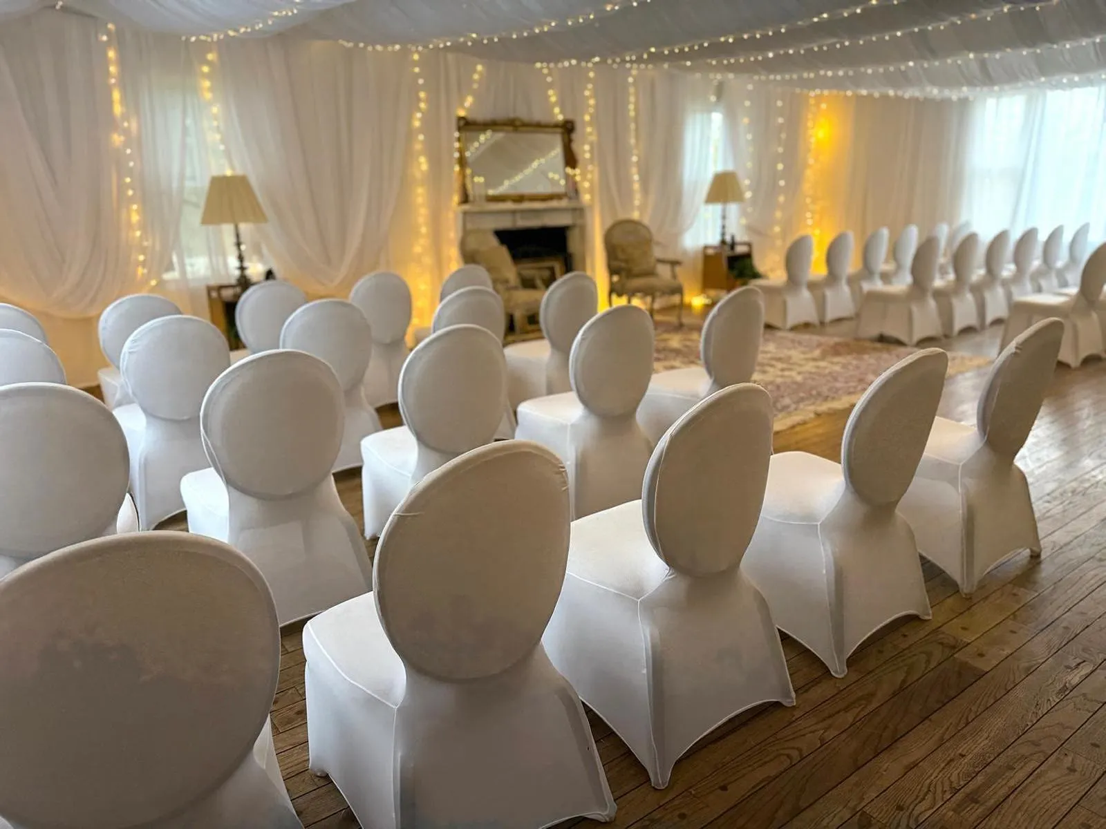 The farmhouse parlor set for a ceremony: white chairs facing a stone fireplace and gilt mirror, framed by draped walls and fairy lights.