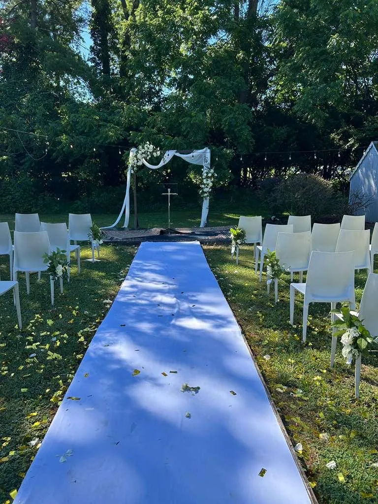 An aisle runner leading through rows of white chairs to a draped arch under mature trees, confetti scattered on the grass.