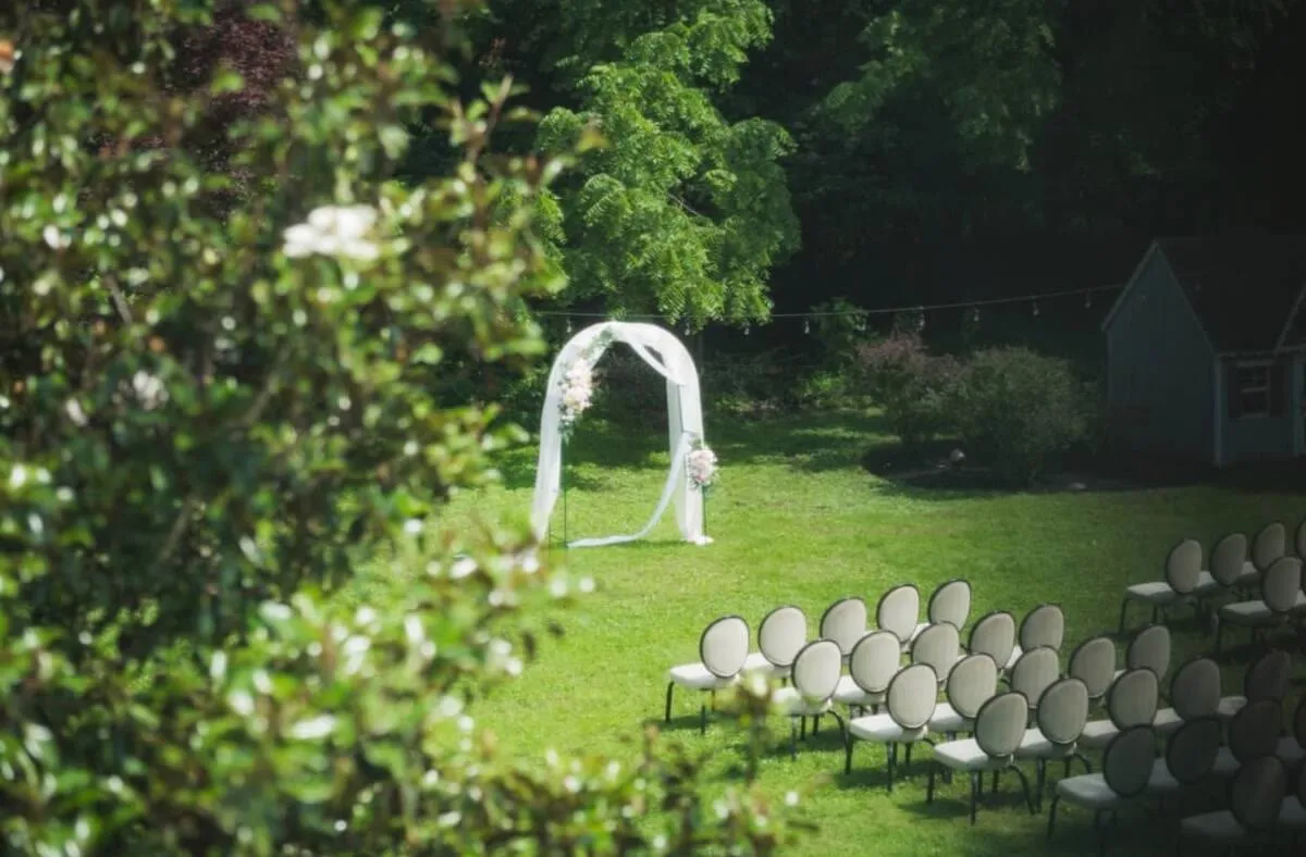 A lawn ceremony seen through foliage — a white draped arch on the grass with rows of chairs set out in front of it.