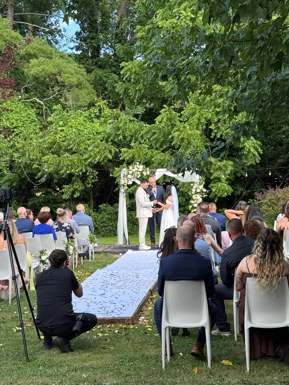 A couple stand with an officiant beneath a white floral arch on the lawn, guests seated on either side of a petal-strewn aisle.