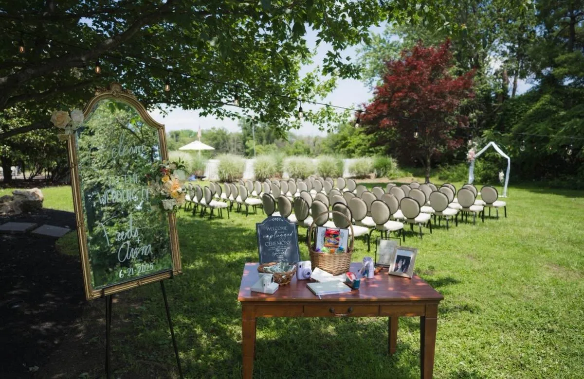 An outdoor ceremony on the lawn with a welcome table and a gold-framed mirror sign at the head of the aisle.