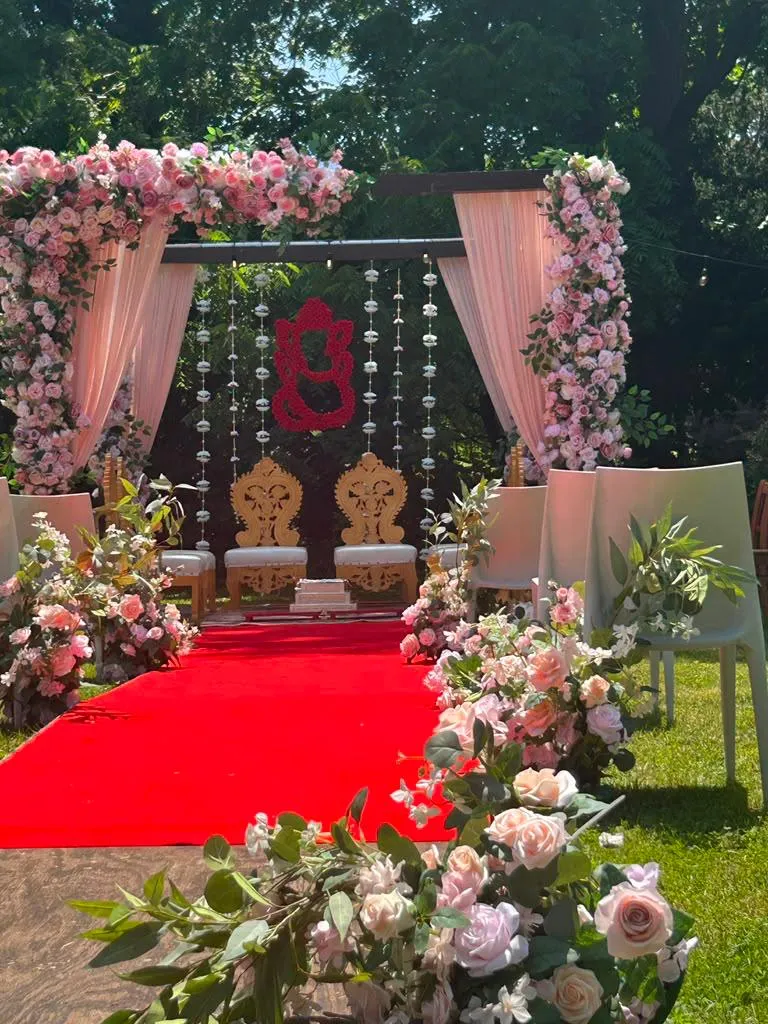 An outdoor ceremony structure on the lawn, hung with blush and pink roses and pale drapery, a red aisle runner leading to two carved gold chairs.